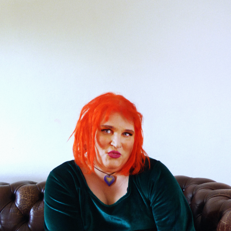 A photograph of Joanna Jones from Saint Ergo. Joanna is sitting on a brown learher chesterfield couch, with a textured white wall behind She has bright Orange Hair, and wearing an Emerald Green Velvet dress. She is pouting with a cheeky smile and looking straight at the camera.